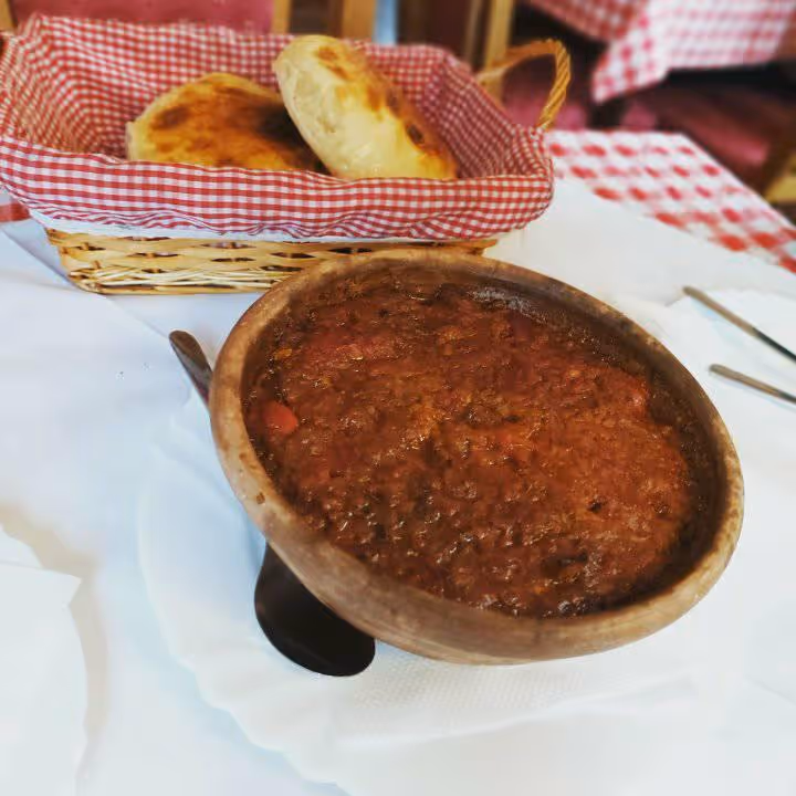 Bowl of thick red stew served on a white plate with a spoon and basket of bread in a red gingham cloth on a restaurant table.