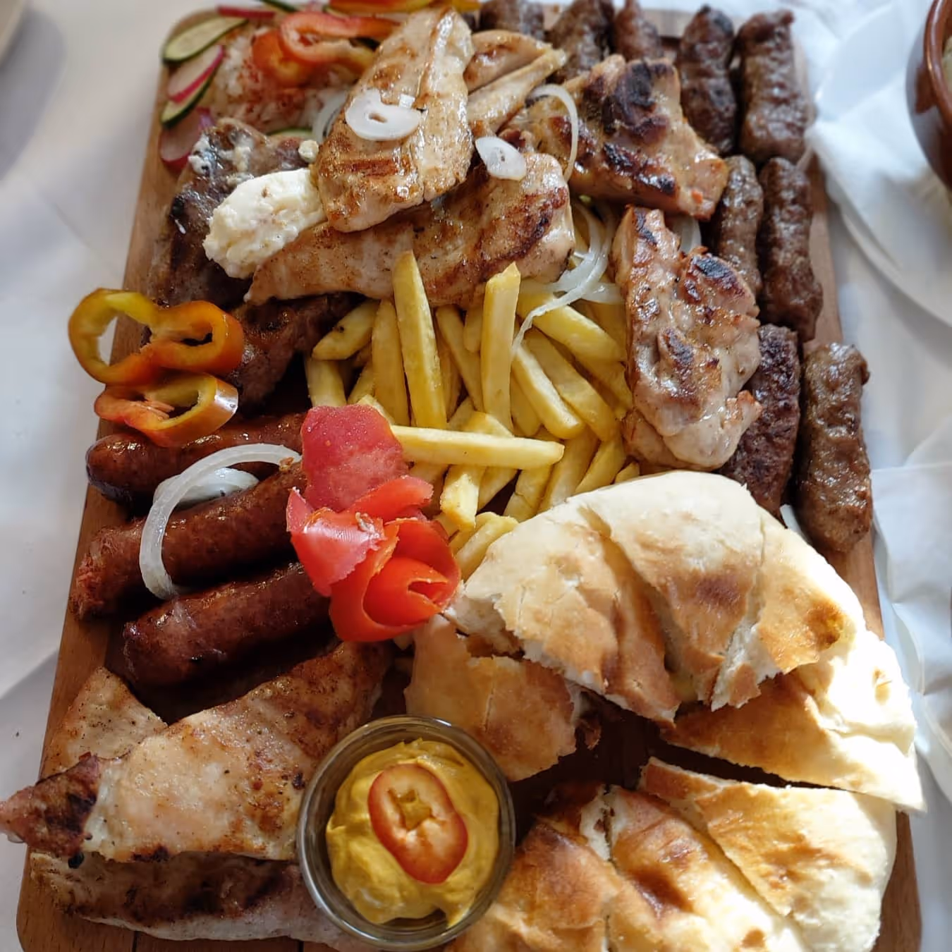Wooden platter with grilled chicken, sausages, meat skewers, French fries, pita bread, mustard sauce, and sliced vegetables.