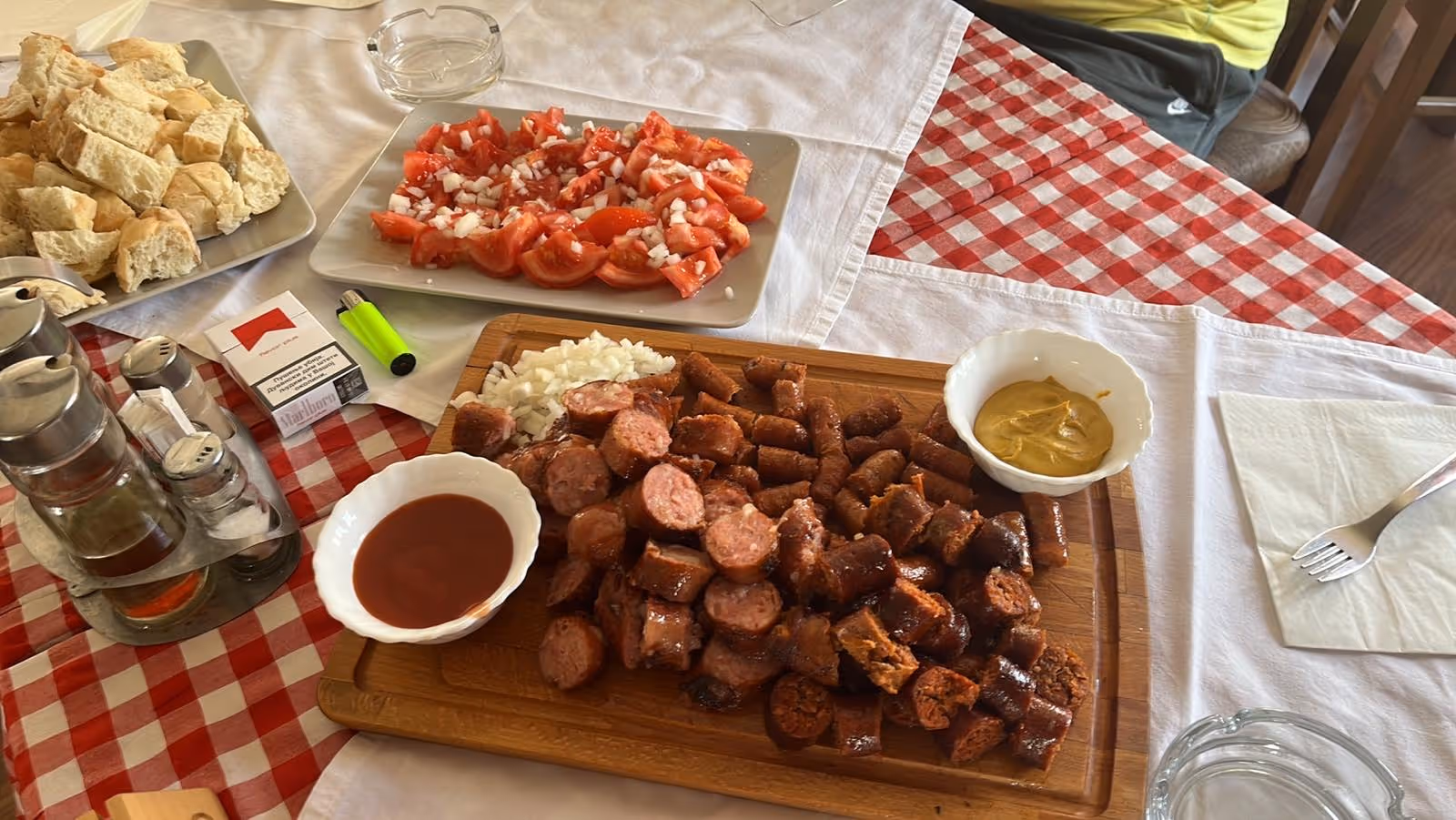 Wooden board with sliced grilled sausages, chopped onions, mustard and ketchup bowls, a plate of chopped tomatoes with onions, and chopped bread on a red and white checkered tablecloth.